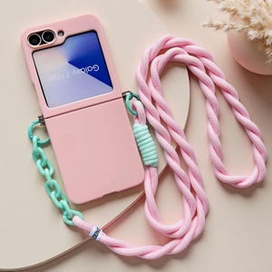 Puede incluir: Una funda de teléfono rosa claro para Galaxy Z Flip6 con una correa larga de cuerda rosa trenzada y un eslabón de cadena de plástico verde menta. La funda tiene un recorte para las lentes de la cámara y una pantalla con el texto Galaxy Z Flip6.