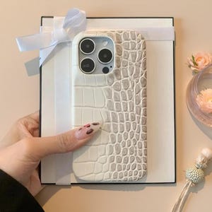 May include: A rectangular white gift box with a white ribbon bow, featuring a phone case with a textured, two-tone crocodile skin pattern in cream and light grey. The case has a cutout for a triple-lens camera system on the upper left side.
