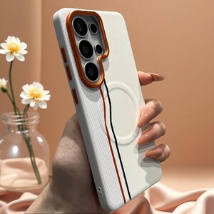 Puede incluir: Una funda de smartphone blanca texturizada con un marco de lente de cámara naranja prominente, un anillo de carga magnético circular en la parte posterior y dos líneas verticales finas en rojo y negro a lo largo del lateral.