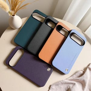 Puede incluir: Una colección de cinco fundas rectangulares para teléfonos móviles en azul verdoso oscuro, negro, naranja, azul claro y morado oscuro. Cada funda presenta un gran recorte para la cámara en la parte superior y un pequeño acento cuadrado metálico con el texto SKIN.