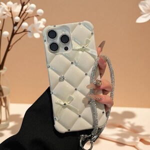May include: A cream-colored, quilted-texture phone case featuring a diamond pattern, small cream bows, and embedded rhinestones. The case includes a matching rhinestone-encrusted wrist strap and a decorative border around the camera lenses.