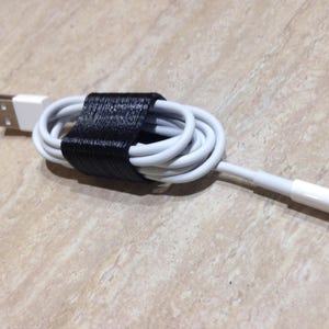 May include: A white charging cable coiled and secured with a black textured cable tie. The cable features a standard USB-A connector on one end and a lightning connector on the other, with a smooth white plastic casing on both ends of the cord.