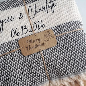 May include: A folded black and white woven blanket with fringe edges, personalized with the text Kaycee & Charlotte and the date 06.13.2026. A brown rectangular tag tied with twine features the text Merry Christmas and a Santa icon.
