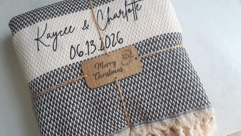 May include: A folded black and white woven blanket with fringe edges, personalized with the text Kaycee & Charlotte and the date 06.13.2026. A brown rectangular tag tied with twine features the text Merry Christmas and a Santa icon.