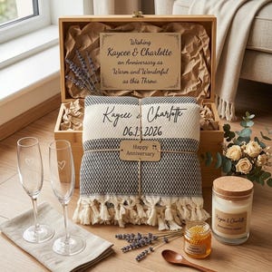 May include: A wooden gift box containing a black and white woven throw blanket with fringe, personalized with names and a date. Includes two champagne flutes with heart etchings, a candle, a jar of honey, a wooden spoon, and dried lavender.