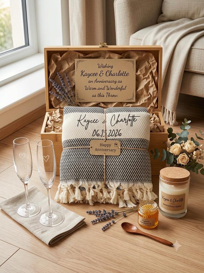 May include: A wooden gift box containing a black and white woven throw blanket with fringe, personalized with names and a date. Includes two champagne flutes with heart etchings, a candle, a jar of honey, a wooden spoon, and dried lavender.