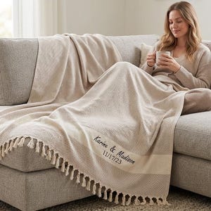 May include: A beige waffle-knit throw blanket with fringe trim on the edges. The blanket features dark embroidery with the text Karine & Madison and the date 11/17/23. The fabric has a textured grid pattern throughout the entire surface area.