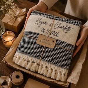 May include: A folded black and cream woven throw blanket with fringe, personalised with the text Kaycee & Charlotte and the date 06.13.2026. A brown paper tag tied with twine reads Happy Anniversary with a small icon of two wine glasses.