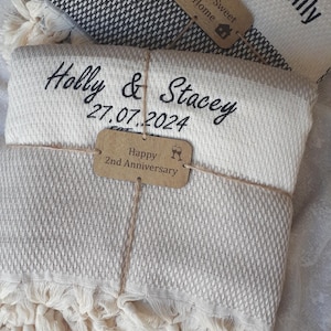 May include: Two folded woven blankets with fringe edges. The top blanket is black and white with text reading The Mose Family Est 2006 and a tag saying Home Sweet Home. The bottom blanket is cream and beige with text Holly & Stacey 27.07.2024.