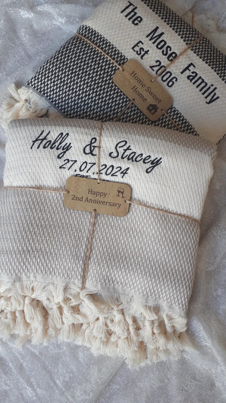 May include: Two folded woven blankets with fringe edges. The top blanket is black and white with text reading The Mose Family Est 2006 and a tag saying Home Sweet Home. The bottom blanket is cream and beige with text Holly & Stacey 27.07.2024.