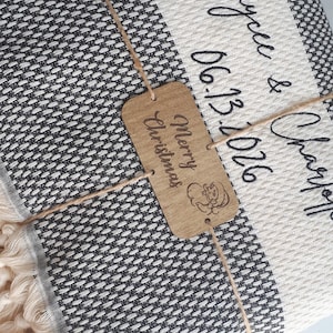 May include: A folded black and white woven throw blanket with fringe, personalized with the text Kaycee & Charles and the date 06.13.2026. A rectangular wooden tag tied with twine features the text Merry Christmas and a small Santa icon.