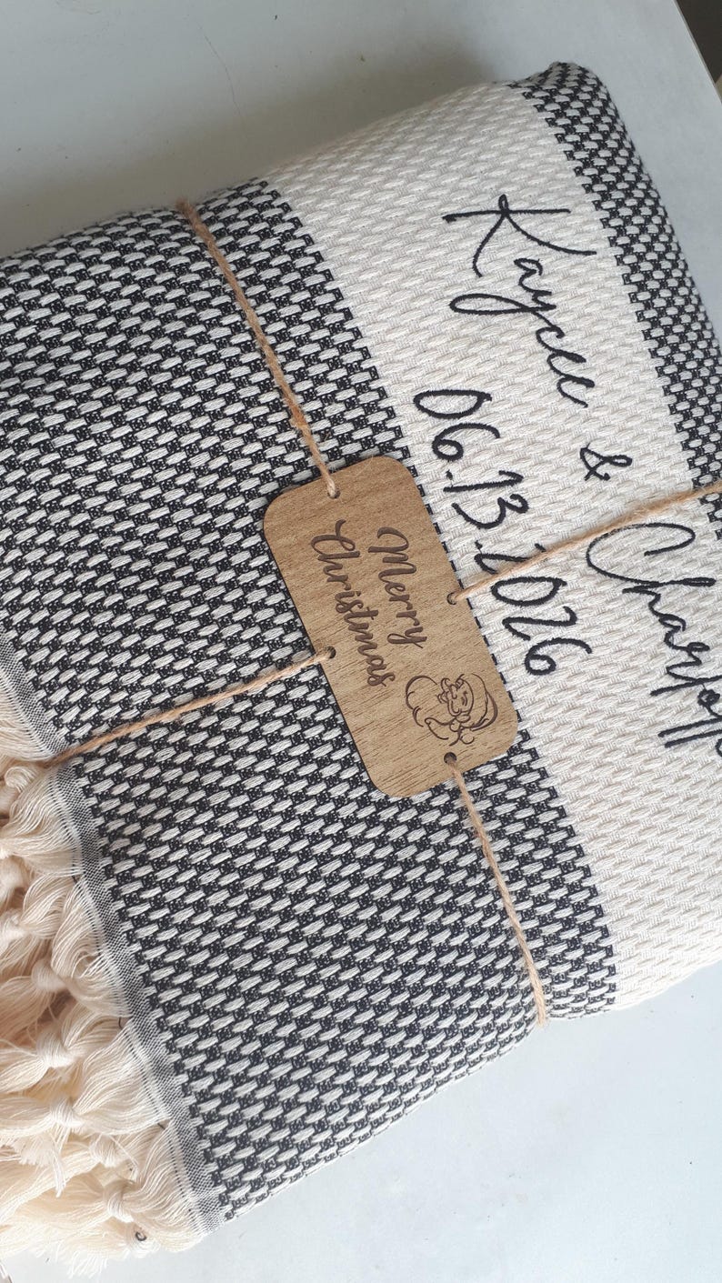 May include: A folded black and white woven throw blanket with fringe, personalized with the text Kaycee & Charles and the date 06.13.2026. A rectangular wooden tag tied with twine features the text Merry Christmas and a small Santa icon.