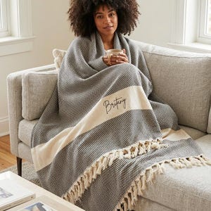 May include: A black and white woven throw blanket featuring a textured pattern, a wide cream-colored horizontal stripe across the center, and decorative fringe along the bottom edge. The name Britany is embroidered in black script on the stripe.