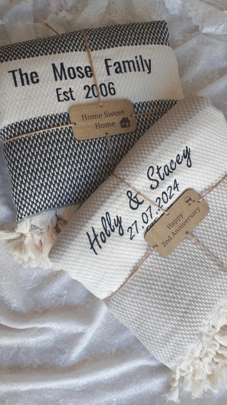 May include: Two folded woven blankets with fringe edges. One blanket is black and white with text reading The Mose Family Est 2006 and a wooden tag saying Home Sweet Home. The other is cream with text Holly & Stacey 27.07.2024 and a tag Happy 2nd Anniversary.