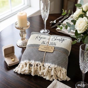 May include: A folded woven throw blanket with cream-colored tassels, featuring the black text Kaycee & Charlotte and 06.13.2026, wrapped with a tan tag reading Happy Anniversary, placed on a dark table with a candle, champagne flute, and locket.