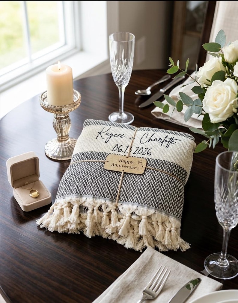 May include: A folded woven throw blanket with cream-colored tassels, featuring the black text Kaycee & Charlotte and 06.13.2026, wrapped with a tan tag reading Happy Anniversary, placed on a dark table with a candle, champagne flute, and locket.