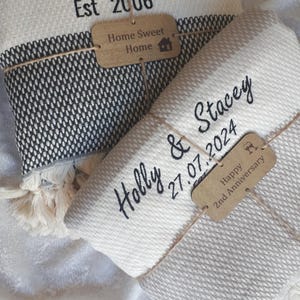 May include: Two folded woven blankets with fringe edges. One blanket is black and white with text reading The Mose Family Est 2006 and a wooden tag saying Home Sweet Home. The other is cream with text Holly & Stacey 27.07.2024 and a tag Happy 2nd Anniversary.