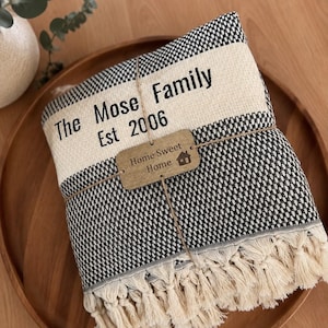 May include: A folded black and white woven throw blanket with cream-colored fringe. The blanket features a cream-colored band printed with the text The Mose Family Est 2006 and a small wooden tag tied with twine reading Home Sweet Home.