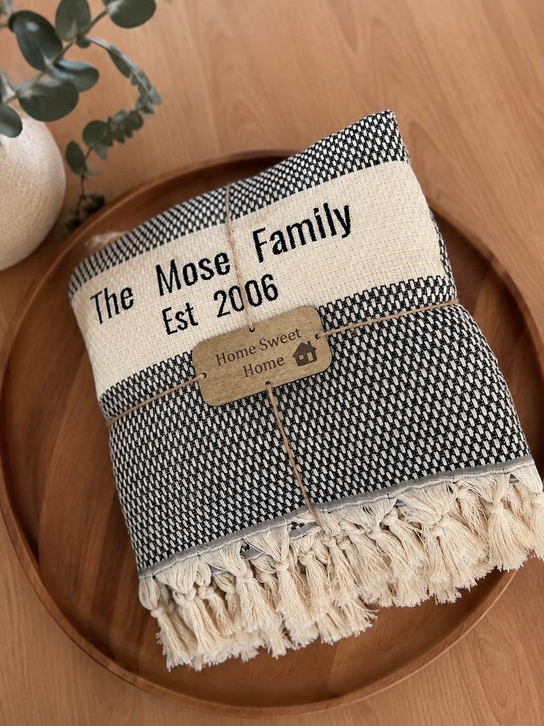 May include: A folded black and white woven throw blanket with cream-colored fringe. The blanket features a cream-colored band printed with the text The Mose Family Est 2006 and a small wooden tag tied with twine reading Home Sweet Home.