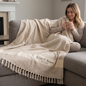 May include: A beige throw blanket with a textured waffle weave pattern and fringe trim on the edges. The blanket features custom embroidery in dark thread that reads Karine & Madison and the date 11/17/23 on a smooth horizontal band.