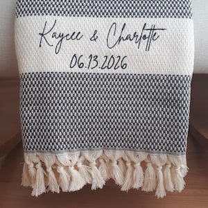 May include: A black and white woven throw blanket featuring a textured pattern and a wide horizontal cream-colored stripe. The text "Kaycee & Charlotte" and "06.13.2026" is embroidered in black script. The bottom edge has cream-colored fringe tassels.