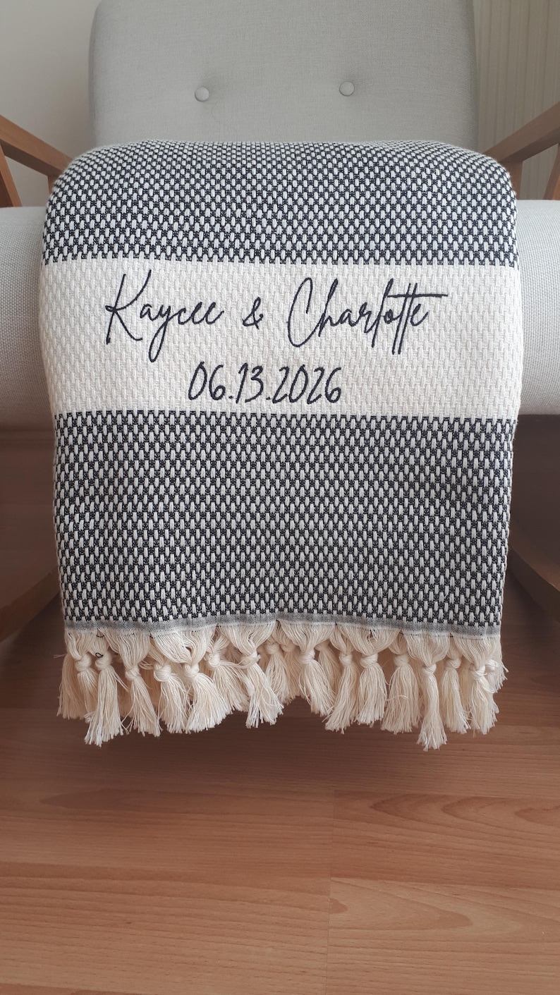 May include: A black and white woven throw blanket featuring a textured pattern and a wide horizontal cream-colored stripe. The text "Kaycee & Charlotte" and "06.13.2026" is embroidered in black script. The bottom edge has cream-colored fringe tassels.