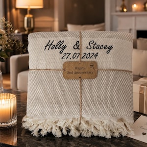 May include: A folded beige woven throw blanket with cream-colored fringe, embroidered with the black text Holly & Stacey and 27.07.2024. A brown rectangular tag tied with twine reads Happy 2nd Anniversary with a small wine glass icon.