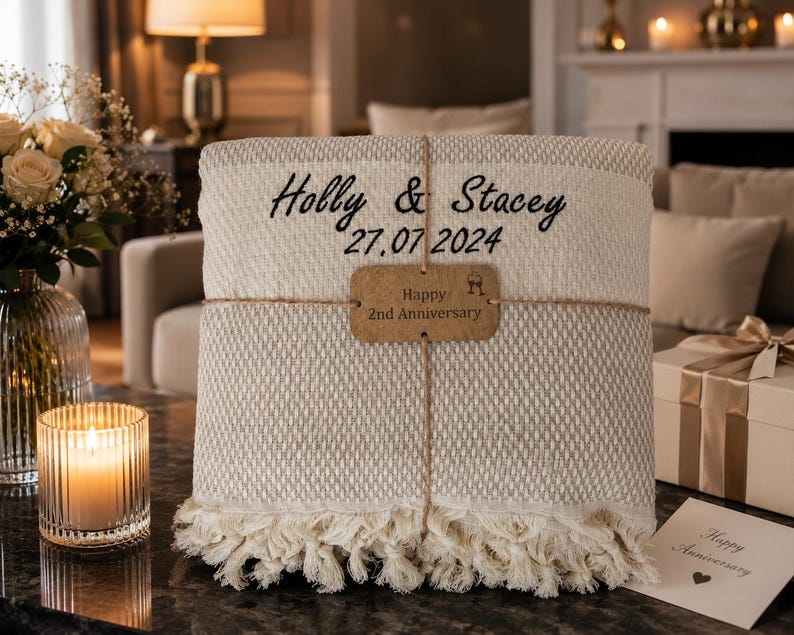 May include: A folded beige woven throw blanket with cream-colored fringe, embroidered with the black text Holly & Stacey and 27.07.2024. A brown rectangular tag tied with twine reads Happy 2nd Anniversary with a small wine glass icon.