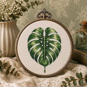 May include: A circular embroidery hoop featuring a cross-stitch design of a single large monstera leaf in shades of green. The leaf is centered on a white fabric background. The hoop has an ornate, antique bronze-colored metal frame.