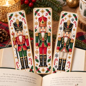 Puede incluir: Tres marcapáginas rectangulares de punto de cruz con figuras de cascanueces en uniformes rojos, verdes y negros. Cada marcapáginas está decorado con hojas de acebo, bayas rojas, estrellas doradas y bastones de caramelo sobre tela blanca.