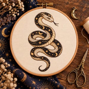 May include: A circular wooden embroidery hoop containing a cross-stitch design of a coiled snake on cream-coloured fabric. The snake features a black and gold pattern with celestial motifs including stars and crescent moons across its body.