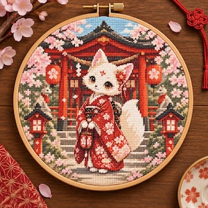 May include: A circular wooden embroidery hoop containing a cross-stitch design of a white fox wearing a red kimono with cherry blossom patterns. The background features a red torii gate, stone lanterns, and pink cherry blossom trees.