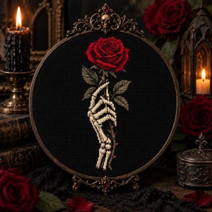 Puede incluir: Un bastidor de bordado circular con un diseño de punto de cruz sobre tela negra. El diseño muestra una mano esquelética sosteniendo una rosa roja vibrante con hojas verdes y un tallo espinoso, enmarcado por un borde metálico ornamentado.
