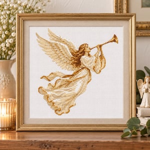 May include: A framed cross-stitch embroidery featuring an angel with large wings, wearing a flowing gown, and playing a golden trumpet. The design uses shades of gold, cream, and white thread against a white fabric background, encased in a gold frame.