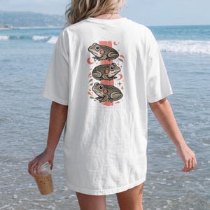 Könnte beinhalten: Ein weißes Kurzarm-T-Shirt mit einer vertikalen Grafik auf der Rückseite. Das Design zeigt drei übereinander angeordnete Illustrationen von gefleckten Fröschen in Braun- und Grautönen, akzentuiert durch kleine rote Kreise, Mondsicheln und Sterne.