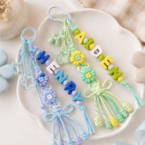 May include: Two macrame keychains featuring knotted cord, flower-shaped accents, and star-shaped charms. One keychain is blue with the name HENRY in block letters, and the other is green with the name AIDEN in block letters, both with clips.