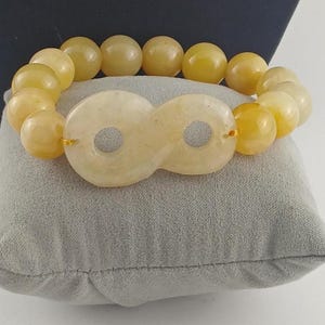 May include: A bracelet composed of smooth, round, pale yellow beads and a central infinity-shaped charm. The beads are uniform in size and color, creating a continuous loop. The central piece features two circular cutouts, maintaining the same light yellow tone as the beads, and is strung together with a thin, flexible cord.