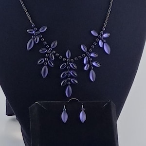Peut inclure: Un ensemble collier et boucles d'oreilles composé de pierres violettes en forme de marquise disposées en motif de feuilles sur une chaîne en métal sombre. Le bijou est présenté avec une boîte bleue et blanche marquée AVON, incluant des boucles.