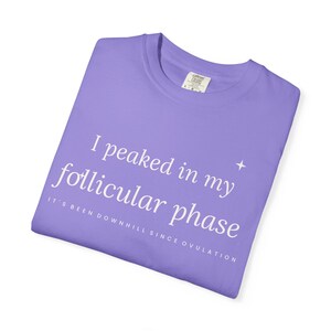 Pode incluir: Uma t-shirt roxo-claro com texto branco que diz "I peaked in my follicular phase" seguido de "IT'S BEEN DOWNHILL SINCE OVULATION" em letras maiúsculas menores, com um pequeno ícone de estrela de quatro pontas à direita do texto.