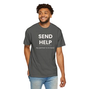 Può includere: Una t-shirt a maniche corte grigio scuro con il testo bianco "SEND HELP" in un carattere sans-serif in grassetto, con una sottile linea orizzontale sotto e il testo bianco più piccolo "my partner is in luteal" stampato sotto.