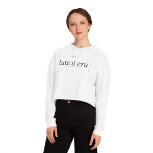 Puede incluir: Una sudadera corta blanca de manga larga con capucha y cordón, que presenta el texto negro "IN MY luteal era" impreso en el pecho junto con pequeños detalles de estrellas, combinada con pantalones negros.
