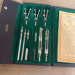 May include: A vintage Dietzgen drafting instrument set in a black folding case with a green velvet interior. The set includes various silver-toned metal compasses, dividers, and drawing tools, alongside a paper Dietzgen Lifetime Service Policy card.