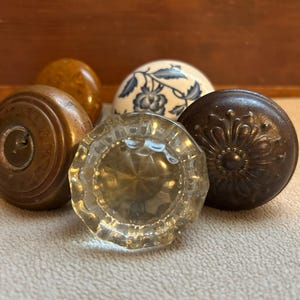 May include: A collection of five assorted door knobs including a clear faceted glass knob, a dark brown metal knob with a floral embossed pattern, a smooth brown wooden knob, a plain amber-colored knob, and a white ceramic knob with blue floral designs.