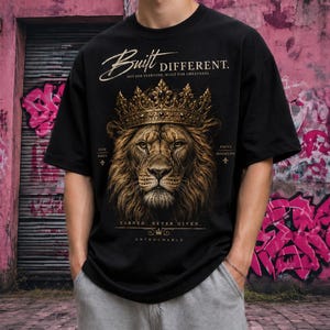 Puede incluir: Una camiseta negra de manga corta con un gráfico de un león con una corona dorada. El texto dice "Built DIFFERENT. NOT FOR EVERYONE, BUILT FOR GREATNESS." con "EXTD MMXV", "FOCUS DISCIPLINE", "EARNED, NEVER GIVEN." y "UNTOUCHABLE" impresos.