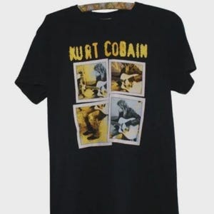 May include: A black short-sleeve t-shirt featuring the text KURT COBAIN in yellow letters at the top. Below the text are four rectangular photo-style prints arranged in a grid, each depicting a person playing an acoustic guitar in various poses.