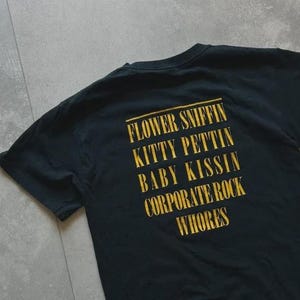 May include: A black short-sleeved t-shirt featuring yellow text printed on the back. The text reads: FLOWER SNIFFIN, KITTY PETTIN, BABY KISSIN, CORPORATE ROCK, WHORES. The shirt has a crew neckline and is made of a solid black fabric.