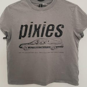 Puede incluir: Una camiseta gris de manga corta con el texto negro 'pixies' sobre un gráfico de un coche fúnebre. Debajo del vehículo, el texto dice 'ON THE GRAVEYARD HILL, SHE'S CALLING OUT HER CURSE'. La camiseta tiene un cuello redondo acanalado y un dobladillo estándar.
