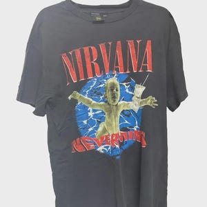 Puede incluir: Una camiseta de manga corta gris carbón con un estampado gráfico de un bebé nadando hacia un billete de dólar. El texto NIRVANA aparece en rojo sobre la imagen y la palabra NEVERMIND está impresa en rojo debajo del gráfico de agua azul.