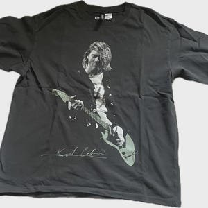 Może przedstawiać: Ciemnoszary t-shirt z krótkim rękawem z czarno-białym nadrukiem graficznym przedstawiającym Kurta Cobaina grającego na gitarze elektrycznej. Projekt zawiera podpis białą czcionką pod gitarą. Koszulka posiada okrągły dekolt i standardowy krój.