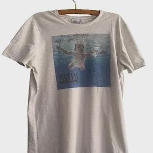 May include: A light gray short-sleeve t-shirt featuring a graphic print of the Nirvana Nevermind album cover, which depicts a baby swimming underwater toward a dollar bill. The word Nirvana is printed in black text at the bottom of the graphic.